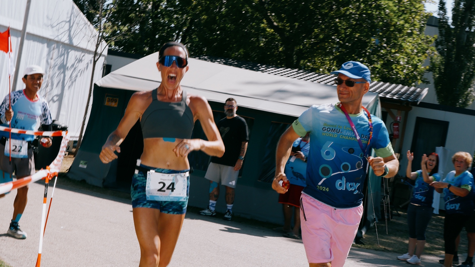 Megan Eckert, wearing sunglasses and bib 24, runs through the EMU finish area as people watch nearby.